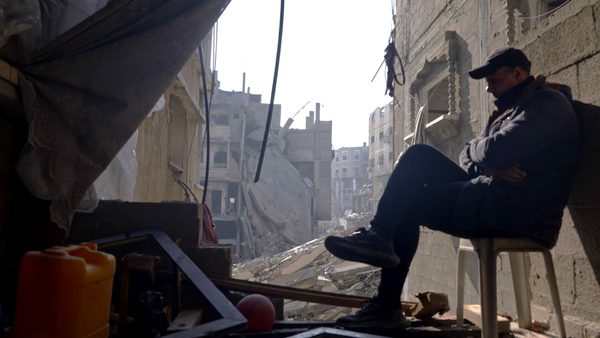 A person with an obscured face sits on a chair inside a heavily damaged building. The background shows collapsed structures and rubble, indicating extensive destruction.