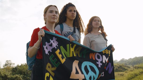 Three individuals stand outdoors holding a banner that reads 'GREENHAM WOMYN 40' with a peace symbol, commemorating the 40th anniversary of the Greenham Common Women's Peace Camp anti-nuclear protest movement.