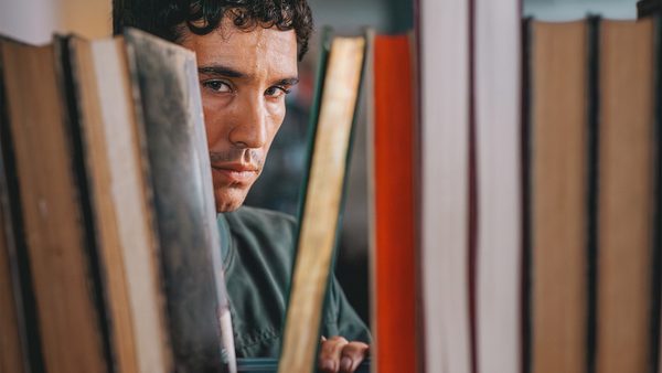 A person peers through a row of vertically arranged books on a shelf, with their face partially obscured, suggesting a moment of curiosity or contemplation in a library or bookstore setting.