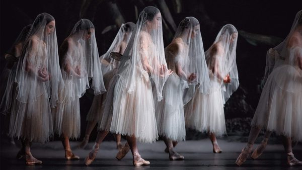 Group of ballet dancers on stage wearing white tutus and veils, performing with graceful arm positions and pointe shoes against a dark theatrical background.