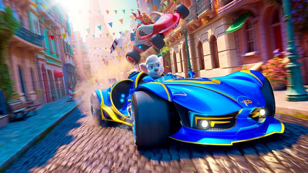 A blue race car with yellow accents speeds through a colourful, narrow street lined with buildings, driven by a white-haired animated character. A red race car with another animated driver is airborne in the background, with festive banners and flags overhead.