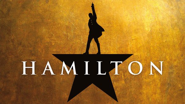 Logo for the musical 'Hamilton' featuring a black silhouette of a person standing on one leg with one arm raised, positioned atop a black star. The background is textured gold, and the word 'HAMILTON' is written in large white capital letters, with the star and silhouette forming part of the letter 'A'.