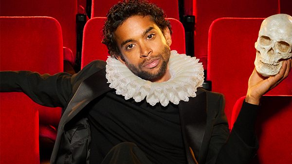 Person in a black outfit with a white ruffled collar sitting in a theater with red seats, holding a skull in their right hand; evoking a scene from Shakespeare's 'Hamlet'.