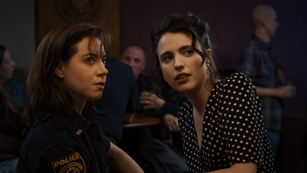 Two people conversing, one in a police uniform and the other in a black dress with white polka dots, with several blurred individuals in the background.