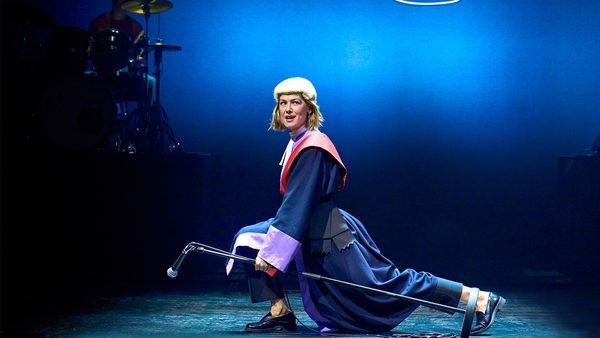 A woman dressed in a legal wig and cloak. She is lunging with her right leg forward whilst holding a microphone and microphone stand.