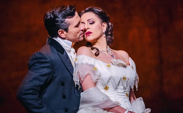 Two people in formal historical attire, one in a dark suit with a white shirt and cravat, the other in an elegant white dress with yellow flowers and lace