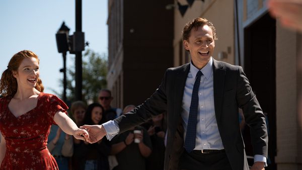 Two people holding hands outdoors, one in a red floral dress and the other in a dark suit with a light blue shirt and dark tie. Blurred figures in the background appear to be taking photos or observing, with buildings and streetlights visible.