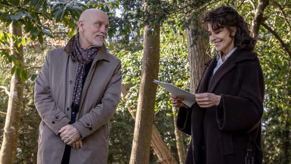 Two individuals stand in a wooded area, one in a light coat and scarf, the other in a dark coat holding a piece of paper, with trees and greenery in the background.