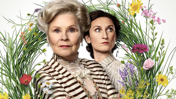Two people standing in front of a circular floral arrangement; one wears a striped outfit with lace, the other a light-coloured shirt.
