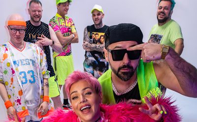 Group of seven adults in bright, retro-inspired outfits posing energetically in a white studio setting.