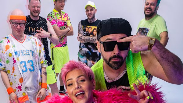 Group of seven adults in bright, retro-inspired outfits posing energetically in a white studio setting.