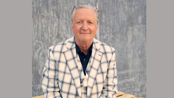Person in a cream plaid suit sitting against a textured grey wall.