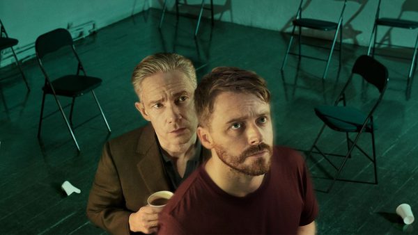 Room with scattered folding chairs and two individuals standing in the foreground, one holding a cup. The wooden floor has some objects on it, including a crumpled piece of paper. Lighting has a greenish tint.