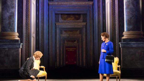 Two people on a stage with an ornate backdrop of tall columns and a long hallway. One person is standing in a bright blue outfit holding a handbag, while the other is kneeling near a gold chair in a striped outfit.