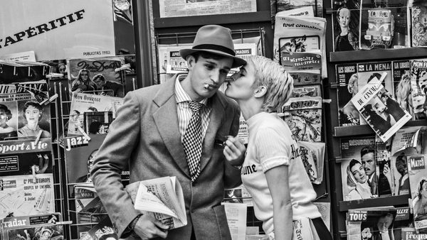 A black-and-white photo showing two people standing in front of a newsstand filled with various magazines and newspapers. One person is wearing a suit, tie, and hat while holding folded newspapers. The other person is leaning toward the suited individual and holding a magazine. The background is densely packed with publications featuring bold headlines and images.