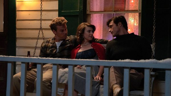 Three people sitting on a porch swing in front of a house with green window frames and pink curtains. The person on the left is holding a bottle, and all appear to be engaged in conversation.