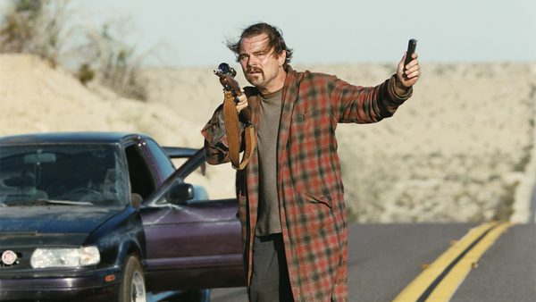 A person with dishevelled hair stands on a deserted road holding a rifle and a handgun, wearing a long plaid coat. An older car with its door open is behind them, set against a dry, hilly landscape.