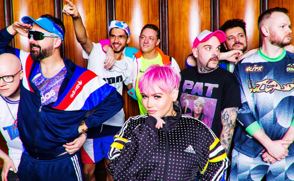 Group of eight people in colorful, retro-style outfits posing together in front of a wooden-paneled wall.