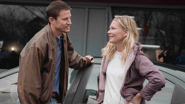 Two people stand beside a car outdoors, conversing. One leans on the car wearing a brown jacket over a blue shirt, while the other stands with hands on hips in a light purple jacket over a white shirt.