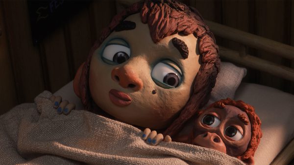 Two claymation characters lying in bed under a blanket. The larger character has blue painted nails. The smaller character, with wide eyes and red hair, peeks out from under the blanket.