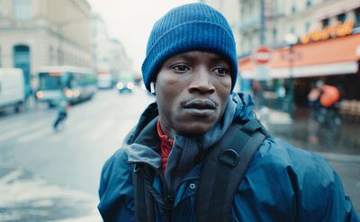Person wearing a blue knit hat and a dark jacket with a backpack, standing on a wet city street with buildings, vehicles, and a café in the background.