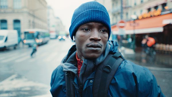Person wearing a blue knit hat and a dark jacket with a backpack, standing on a wet city street with buildings, vehicles, and a café in the background.