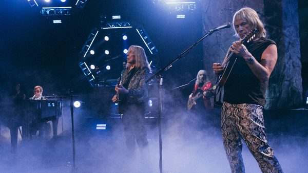 Four musicians perform on a fog-filled stage with dramatic lighting. One plays the keyboard, two play guitars, and one plays bass, all deeply engaged in the live performance.