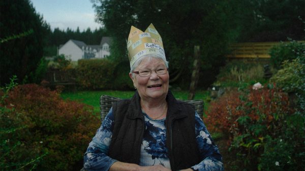 A person sits outdoors in a garden or backyard, wearing a paper crown labelled 'Raffle' with various numbers on it. The background includes trees, bushes, and houses.