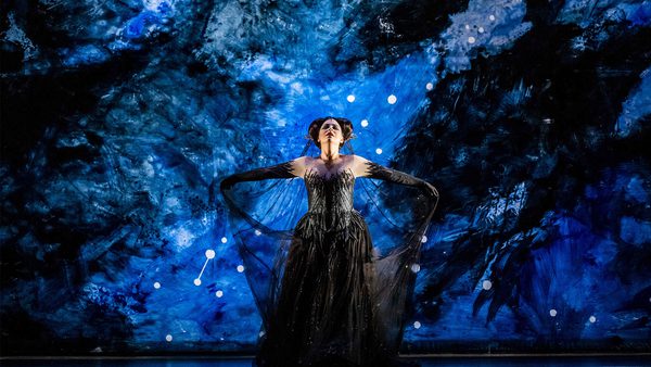 Person standing on a stage with a dramatic blue and black background resembling a night sky or outer space, wearing an elaborate black dress with long sleeves and arms extended outward.