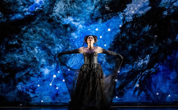 Person standing on a stage with a dramatic blue and black background resembling a night sky or outer space, wearing an elaborate black dress with long sleeves and arms extended outward.