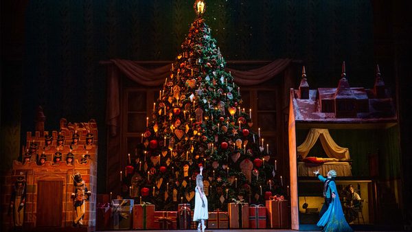 Stage scene from a performance resembling 'The Nutcracker,' featuring a large decorated Christmas tree with ornaments and candles, wrapped presents at its base, toy soldiers near a castle-like structure on the left, and a house-like set on the right with two characters—one in white near the tree and one in blue gesturing toward them.