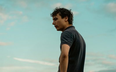 A person standing outdoors under a blue sky with scattered clouds, wearing a dark short-sleeved shirt.