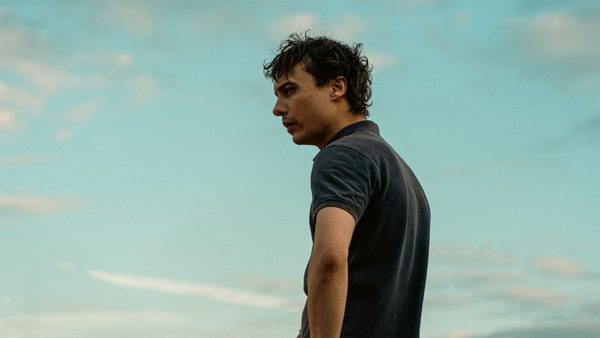 A person standing outdoors under a blue sky with scattered clouds, wearing a dark short-sleeved shirt.