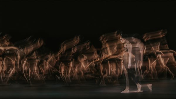 Group of dancers captured in motion with a long exposure effect, creating blurred streaks of light and shadow against a dark background; two figures in the foreground appear more clearly.
