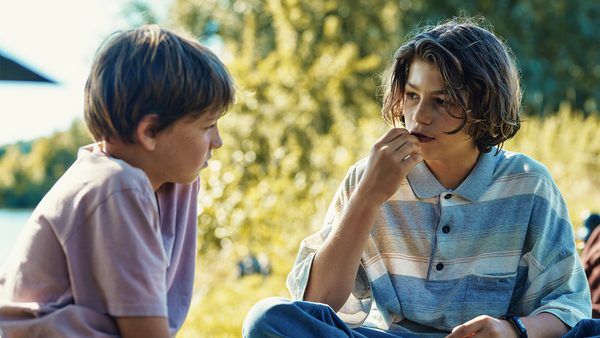 Two individuals sitting outdoors in a natural setting, facing each other and conversing. One wears a light-coloured short-sleeved shirt, the other a striped long-sleeved shirt.