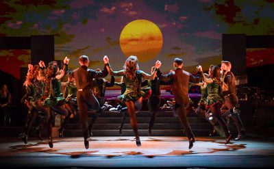 A line of dancers stand in a circle on stage facing away from the audience. The dancers are leaping in the air with arms raised and holding the hands of the dancer next to them.