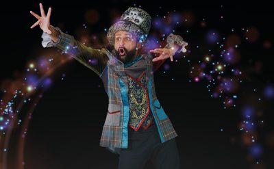 A performer in a colourful, eccentric costume poses with arms outstretched against a dark background. They wear a patterned jacket, ornate gold vest and a large decorative hat with a playing‑card motif. Bright multi-coloured light effects and sparkles surround them, creating a magical, theatrical look.