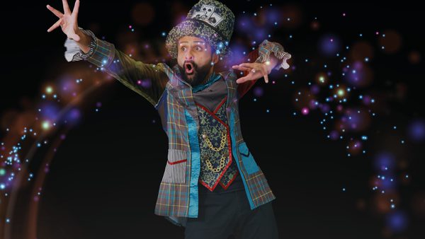 A performer in a colourful, eccentric costume poses with arms outstretched against a dark background. They wear a patterned jacket, ornate gold vest and a large decorative hat with a playing‑card motif. Bright multi-coloured light effects and sparkles surround them, creating a magical, theatrical look.