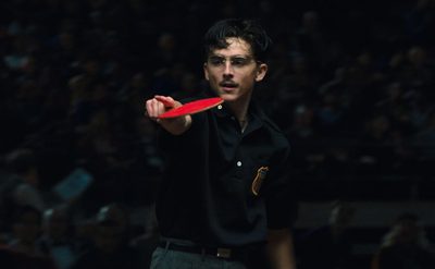 A man with dark curly hair wearing black glasses and a black polo shirt, stands holding a red ping pong bat pointing towards his opponent. There is a seated crowd in the dark behind him.