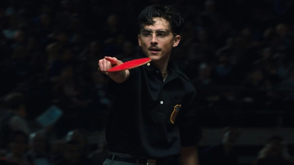 A man with dark curly hair wearing black glasses and a black polo shirt, stands holding a red ping pong bat pointing towards his opponent. There is a seated crowd in the dark behind him.