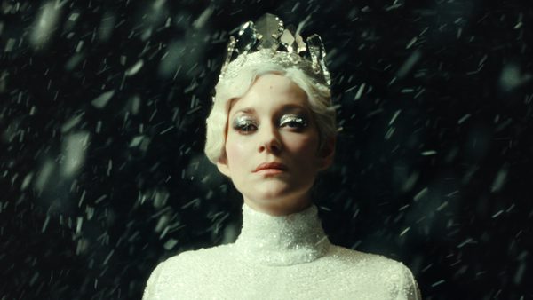 A person stands in falling snow, wearing a shiny white outfit and a crystal-like crown. They have pale blond hair and silver makeup, with a dark background behind them.