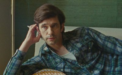 A person lies on a couch wearing a plaid shirt, holding a cigarette and looking thoughtful. There’s a green wall and a lamp in the background.