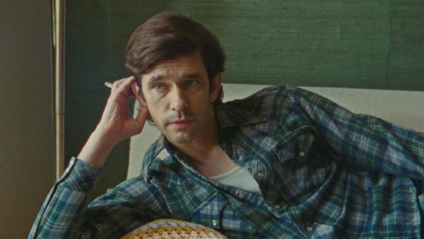 A person lies on a couch wearing a plaid shirt, holding a cigarette and looking thoughtful. There’s a green wall and a lamp in the background.