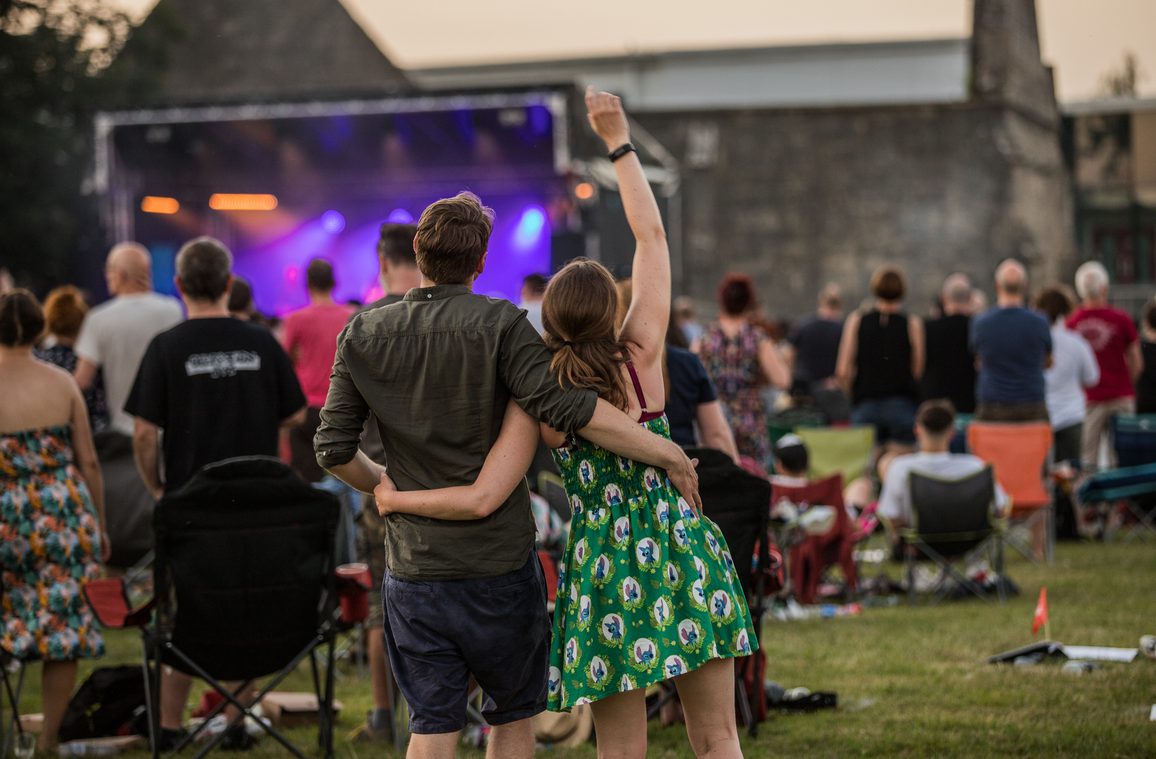 A couple standing arm-in-arm at an outdoor music event, facing a stage with bright purple lights. The crowd is seated on folding chairs and blankets on a grassy field, with a stone building in the background.