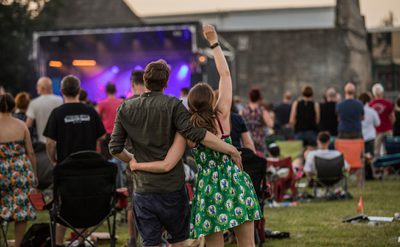 A couple standing arm-in-arm at an outdoor music event, facing a stage with bright purple lights. The crowd is seated on folding chairs and blankets on a grassy field, with a stone building in the background.