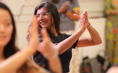 A person with long hair and glasses clapping hands in a group setting, wearing a black shirt. Other people in the background are also clapping, indicating participation in an activity or class.