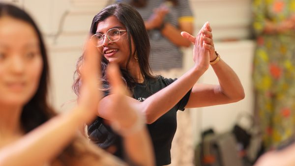 A person with long hair and glasses clapping hands in a group setting, wearing a black shirt. Other people in the background are also clapping, indicating participation in an activity or class.