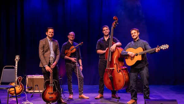A group of four musicians stand on a stage under blue and purple lighting. They hold an archtop guitar, a violin, an upright bass, and an acoustic guitar. A second guitar rests on a chair beside them, with an amp, cables, and pedals on the floor. The scene suggests a live performance or band promo shot.