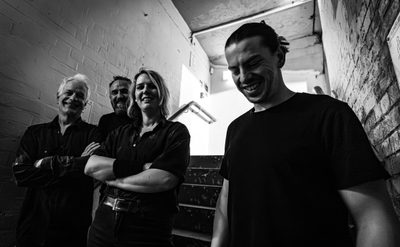 A black and white photo of 4 people, 3 men and 1 woman, standing on some stairs. 3 members of the group stand close to the stairs handrail, they are looking at the camera smiling, the fourth member of the band stands closest to the camera and looks down smiling.