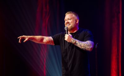 A man wearing a black t-shirt holding a microphone on stage, he is smiling and looking out towards with audience with his hand pointing out in front of him.